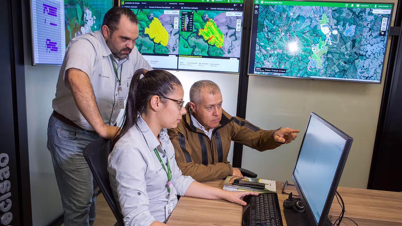 tres pessoas em frente ao computador e atras deles tres tv exibindo mapa de algum local