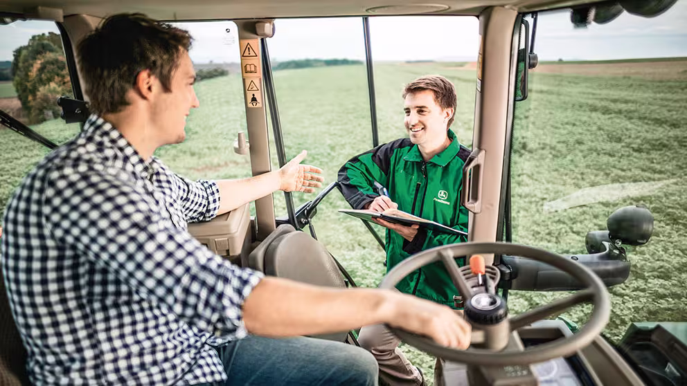foto de dentro de um trator homen com uma mao segurando volante e um tecnico da John Deere na porta com caderno na mao